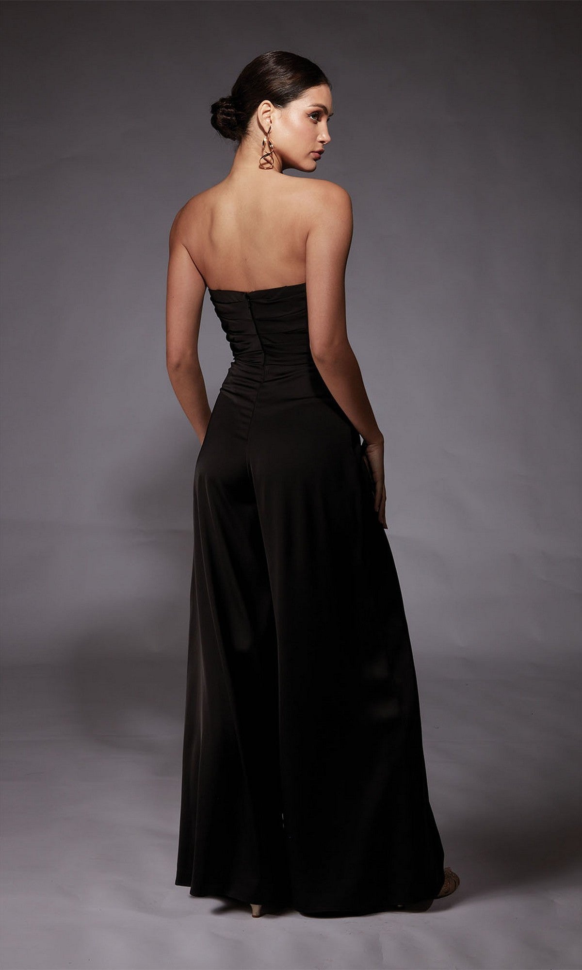Woman stands with her back to the camera in a strapless black Long Formal Jumpsuit: Alyce 70071, featuring wide-leg pants and large gold earrings, set against a plain gray background.